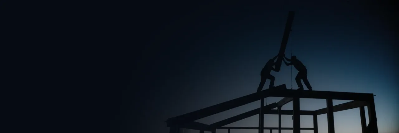Two builders in silhouette raising a steel roof beam into place on an unfinished frame against a deep blue dusk sky